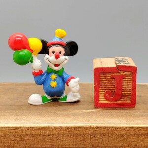 Może przedstawiać: Mała plastikowa figurka klauna Mickey Mouse'a trzymająca trzy balony. Balony są czerwone, ż&oacute;łte i zielone. Klaun ma na sobie niebiesko-ż&oacute;łty kostium z czerwonym nosem i ż&oacute;łtym kapeluszem. Figurka stoi obok drewnianego klocka z namalowaną literą "J".