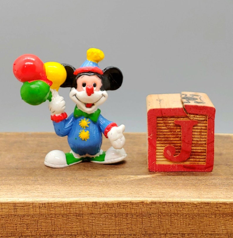 Może przedstawiać: Mała plastikowa figurka klauna Mickey Mouse'a trzymająca trzy balony. Balony są czerwone, ż&oacute;łte i zielone. Klaun ma na sobie niebiesko-ż&oacute;łty kostium z czerwonym nosem i ż&oacute;łtym kapeluszem. Figurka stoi obok drewnianego klocka z namalowaną literą "J".
