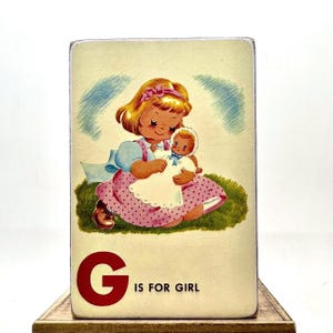 May include: A vintage-style wooden block featuring a cartoon illustration of a girl holding a doll. The girl has blonde hair and wears a blue dress and pink polka-dot skirt. The letter "G" and the phrase "IS FOR GIRL" are printed in red.