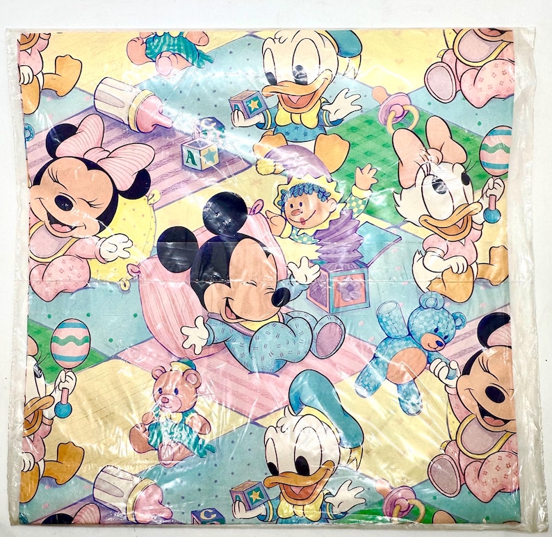 May include: A square sheet of baby-themed wrapping paper featuring cartoon characters Mickey Mouse, Minnie Mouse, Donald Duck, and Daisy Duck. The characters are in pastel colours, surrounded by toys and blocks on a checkered background.