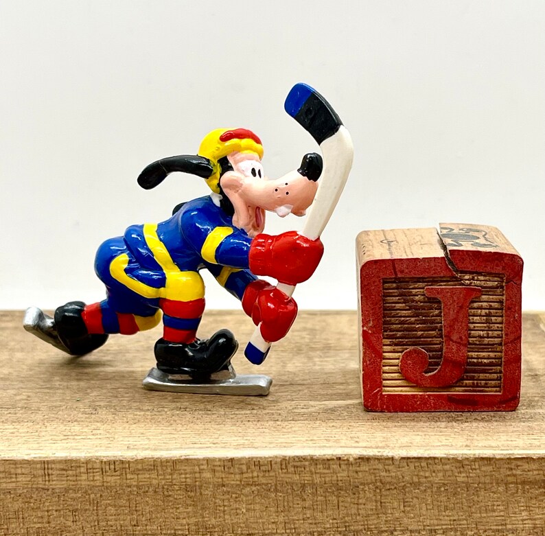May include: A blue, yellow, and red cartoon dog figure holding a hockey stick. The figure is on a wooden surface next to a wooden block with the letter 'J' on it.