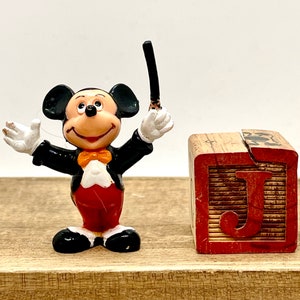 May include: A black and white Mickey Mouse figurine wearing a tuxedo and holding a conductor's baton. The figurine is standing next to a wooden block with the letter 'J' painted on it.