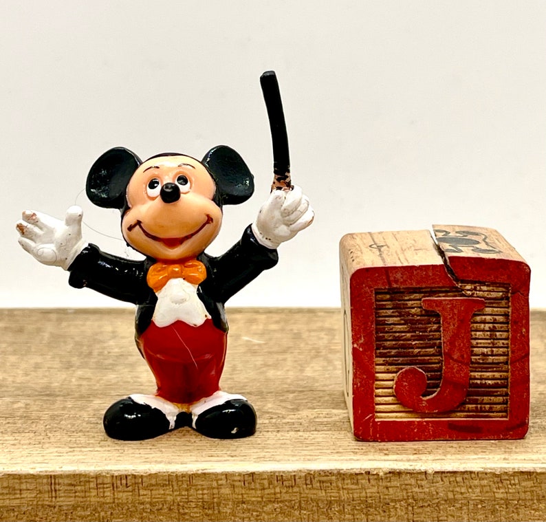 May include: A black and white Mickey Mouse figurine wearing a tuxedo and holding a conductor's baton. The figurine is standing next to a wooden block with the letter 'J' painted on it.