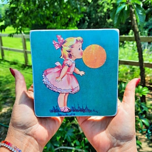 Puede incluir: Un bloque de madera cuadrado con una ilustración vintage de una niña soplando una burbuja. La niña lleva un vestido rosa y un lazo rojo. El fondo es turquesa y la burbuja es naranja.
