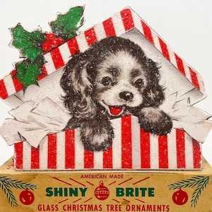 Puede incluir: Una caja de rayas rojas y blancas con un peque&ntilde;o cachorro marr&oacute;n y blanco asom&aacute;ndose por la parte superior. La caja est&aacute; decorada con una rama de acebo verde y bayas. La caja est&aacute; etiquetada con "Shiny Brite Glass Christmas Tree Ornaments" y "American Made".