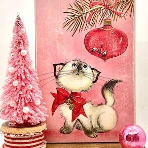 以下が含まれることがあります： ピンクのクリスマスカードに、赤いリボンをつけた白い猫が、松の枝にぶら下がっている赤いオーナメントを見上げています。カードは、ピンクのクリスマスツリーとピンクのオーナメントのある木製の棚の上に置かれています。