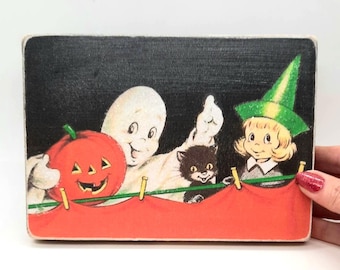 May include: A rectangular wooden sign featuring a vintage-style illustration of a white ghost holding an orange jack-o'-lantern, a small black cat, and a child wearing a green witch hat, all positioned behind a red fabric draped over a green line.
