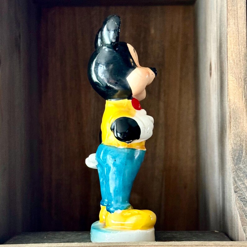 May include: A vintage Mickey Mouse figurine. The figure is painted with black, yellow, blue, white, and flesh-tone colors. Mickey is wearing a yellow jacket, blue pants, and yellow shoes. The figurine is standing on a light blue base.