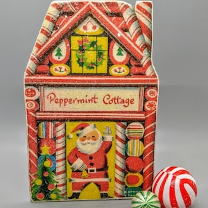 Puede incluir: Un adorno navide&ntilde;o vintage de cart&oacute;n en forma de casita de jengibre con una ilustraci&oacute;n de Pap&aacute; Noel y detalles de bastones de caramelo. El adorno dice "Peppermint Cottage".