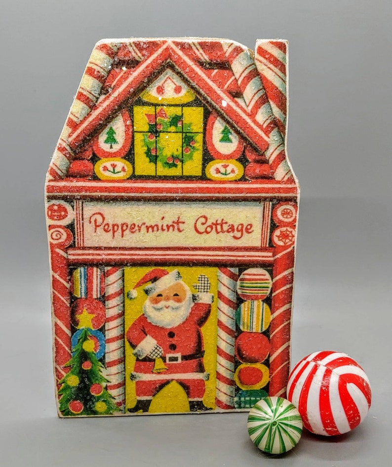 Puede incluir: Un adorno navide&ntilde;o vintage de cart&oacute;n en forma de casita de jengibre con una ilustraci&oacute;n de Pap&aacute; Noel y detalles de bastones de caramelo. El adorno dice "Peppermint Cottage".
