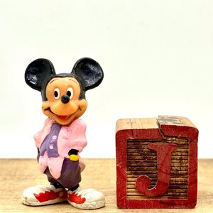 May include: A plastic figurine of Mickey Mouse wearing a pink and purple outfit stands next to a wooden block with the letter "J" painted in red.