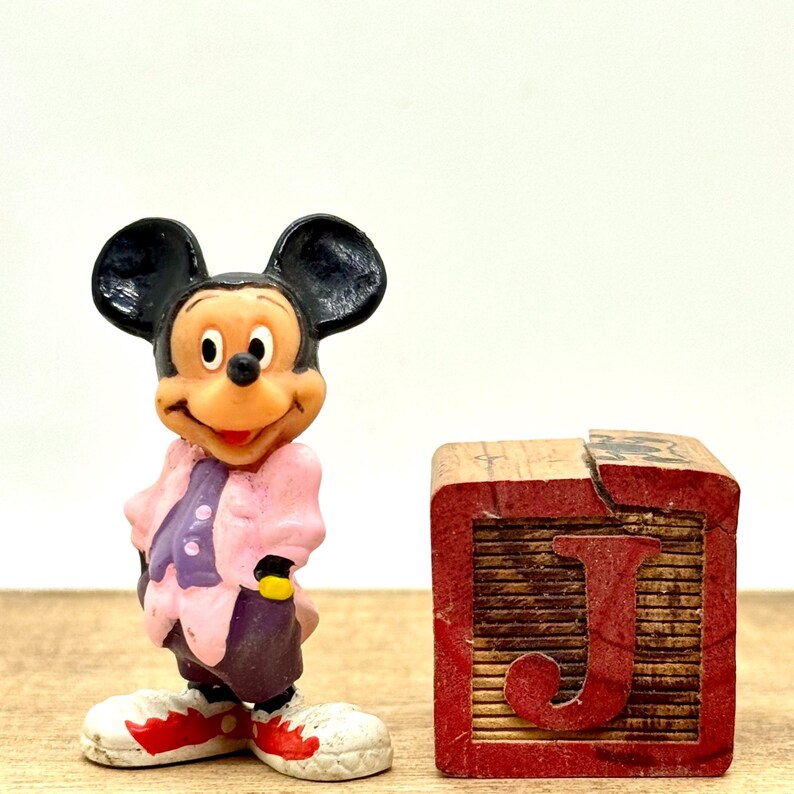 May include: A plastic figurine of Mickey Mouse wearing a pink and purple outfit stands next to a wooden block with the letter "J" painted in red.