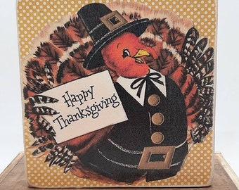 Op de afbeelding: Een houten blok met een vintage illustratie van een kalkoen die een pelgrimhoed draagt en een bord vasthoudt met de tekst "Happy Thanksgiving". De achtergrond is een gouden stippenpatroon.