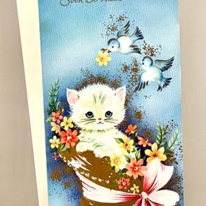 May include: A vintage get-well card with a blue background. It features a white kitten in a basket of flowers, two blue birds, and the text "May You Soon Be Well". A pink bow adorns the basket.