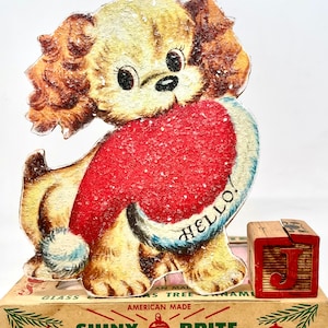 Puede incluir: Un adorno de &aacute;rbol de Navidad vintage con un perro de dibujos animados que lleva un gorro de Pap&aacute; Noel rojo con la palabra "HELLO!" en &eacute;l. El adorno est&aacute; hecho de cart&oacute;n y tiene un acabado brillante. El adorno est&aacute; sentado en una caja que dice "SHINY BRITE GLASS CHRISTMAS TREE ORNAMENTS".