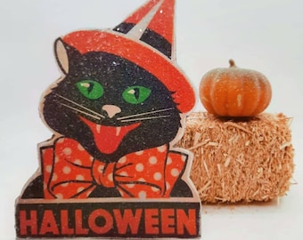 May include: A black cat wearing a red and black witch hat with white polka dots and a red bow tie. The cat is smiling with its tongue sticking out. The cat is standing on a bale of hay with a small orange pumpkin on top. The image says "HALLOWEEN" at the bottom.
