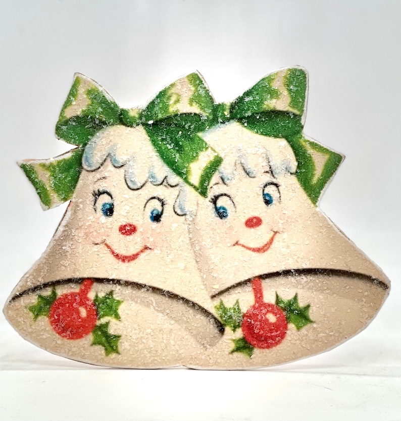 Puede incluir: Dos campanas blancas de Navidad con lazos verdes y detalles de acebo. Las campanas tienen caras sonrientes y est&aacute;n cubiertas de un material blanco brillante.