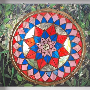 May include: A stained glass window with a circular design. The window features a central starburst pattern in red, blue, and pink, surrounded by a ring of alternating colors. The frame is a warm brown, and the background shows green foliage.