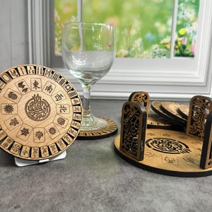 May include: Wooden coasters with a Mahjong theme. The coasters are round with intricate laser-cut designs featuring Chinese characters and symbols. A wine glass sits next to the coasters. A holder with decorative cutouts is also present.