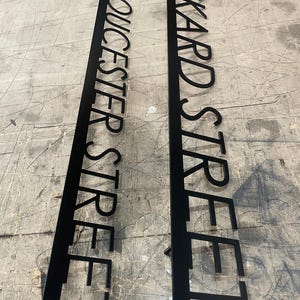 Two long, black metal signs with laser-cut text, one reading "24 GLOUCESTER STREET" and the other "24 RICKARD STREET," lying on a textured gray surface.
