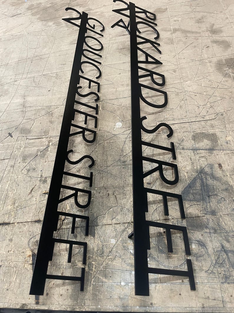 Two long, black metal signs with laser-cut text, one reading "24 GLOUCESTER STREET" and the other "24 RICKARD STREET," lying on a textured gray surface.