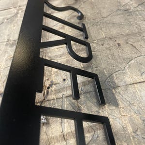 A close-up, side-angle view of the black metal sign, showing the laser-cut letters "STREET" with a slight shadow beneath them on a gray surface.