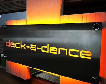 May include: A metal sign with the text "deck-a-dence" in orange lettering, illuminated from behind with warm orange light.