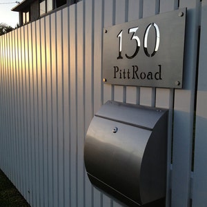 May include: A silver mailbox with a silver house number sign that reads "130 Pitt Road" mounted on a white wooden fence.