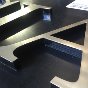 Puede incluir: Primer plano de dos letras de metal plateado, "A" e "I", sobre un fondo negro. Las letras son en relieve y tienen un acabado cepillado.