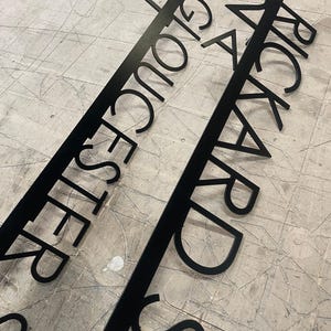 Two black metal signs with laser-cut text, one reading "24 GLOUCESTER STREET" and the other "24 RICKARD STREET," lying on a gray, textured surface.