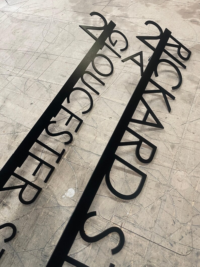 Two black metal signs with laser-cut text, one reading "24 GLOUCESTER STREET" and the other "24 RICKARD STREET," lying on a gray, textured surface.