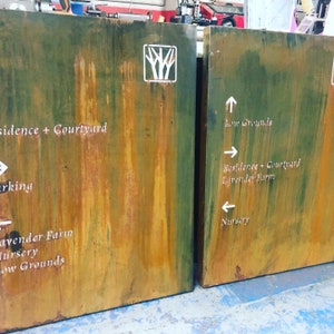May include: Two rusty metal signs with white painted arrows and text. The sign on the left says "Residence + Courtyard", "Parking", "Lavender Farm", "Nursery", and "Low Grounds". The sign on the right says "Low Grounds", "Residence + Courtyard", "Lavender Farm", and "Nursery".