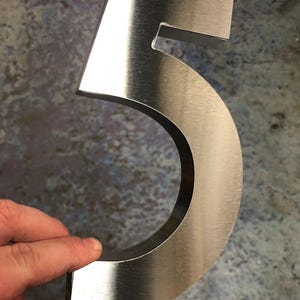 A hand holds a brushed metal number "5", showing its thickness and reflective surface.