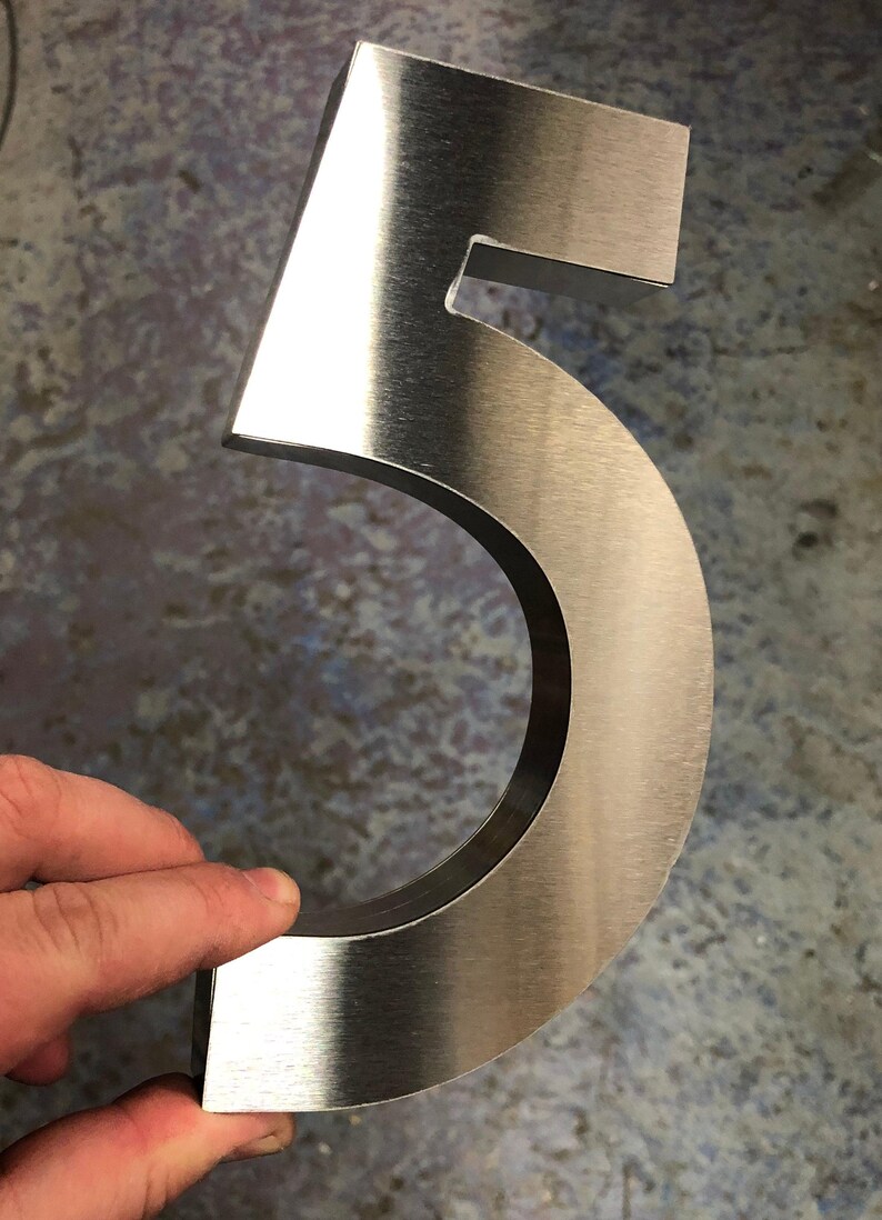 A hand holds a brushed metal number "5", showing its thickness and reflective surface.