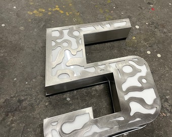 May include: A silver metal number five with a white background and a cutout design. The number is lying on a gray surface.