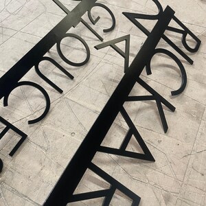 Two black metal signs with laser-cut text, "24 GLOUCESTER STREET" and "24 RICKARD STREET," laid diagonally across a gray, textured surface.