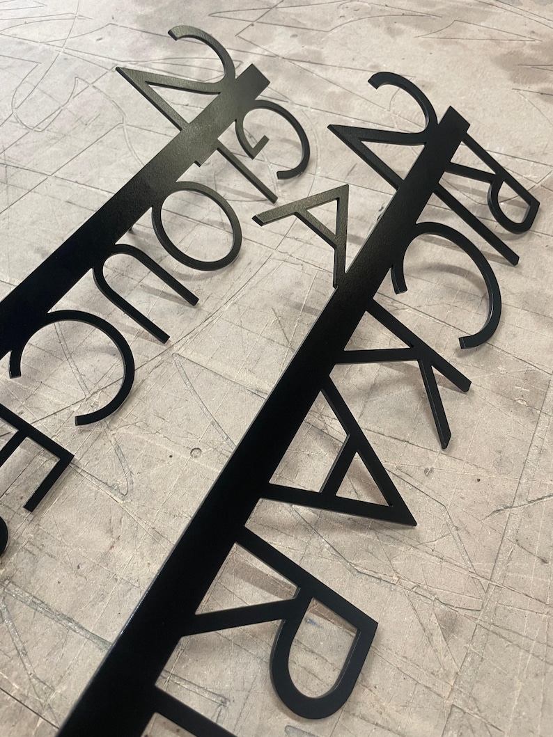 Two black metal signs with laser-cut text, "24 GLOUCESTER STREET" and "24 RICKARD STREET," laid diagonally across a gray, textured surface.
