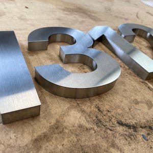 Brushed metal numbers "1312" are shown at an angle on a textured wooden surface, with the "1" and "2" in focus.