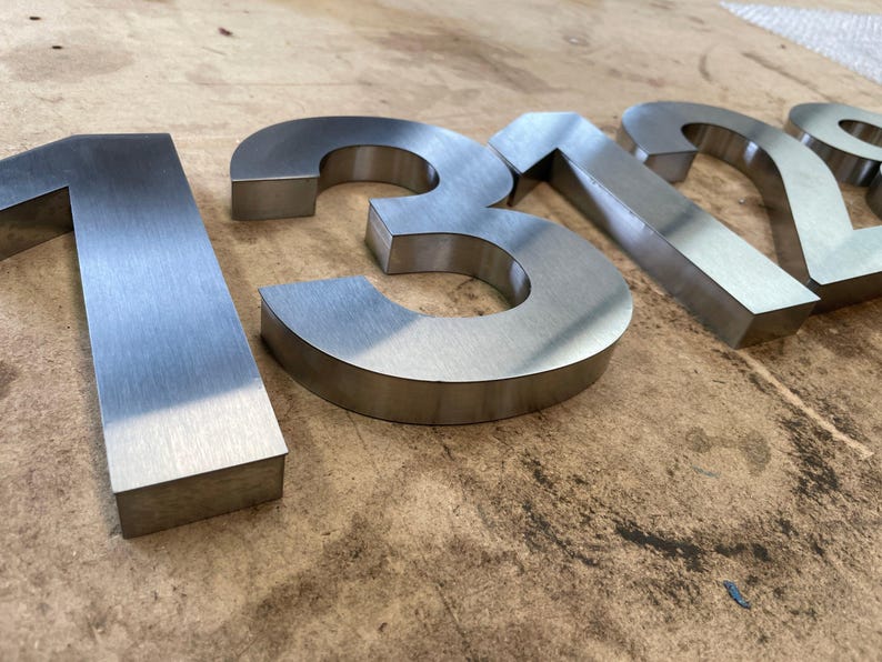 Brushed metal numbers "1312" are shown at an angle on a textured wooden surface, with the "1" and "2" in focus.