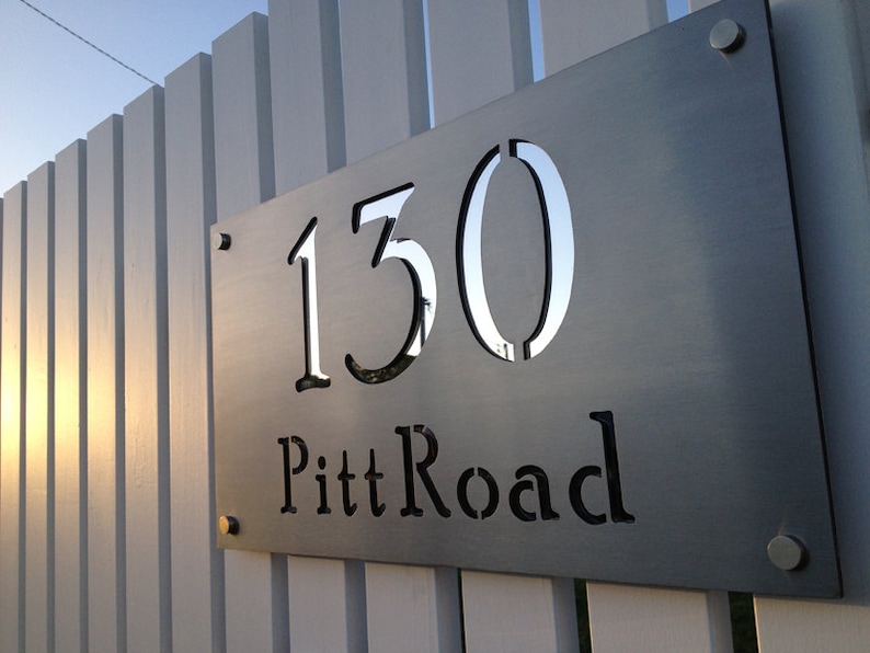 May include: A silver metal house number sign with the number 150 and the street name Pitt Road cut out of the metal. The sign is attached to a white wooden fence.