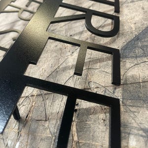A close-up, low-angle view of the black metal sign, showing the laser-cut letters "STREET" with a slightly textured matte finish, on a gray surface.