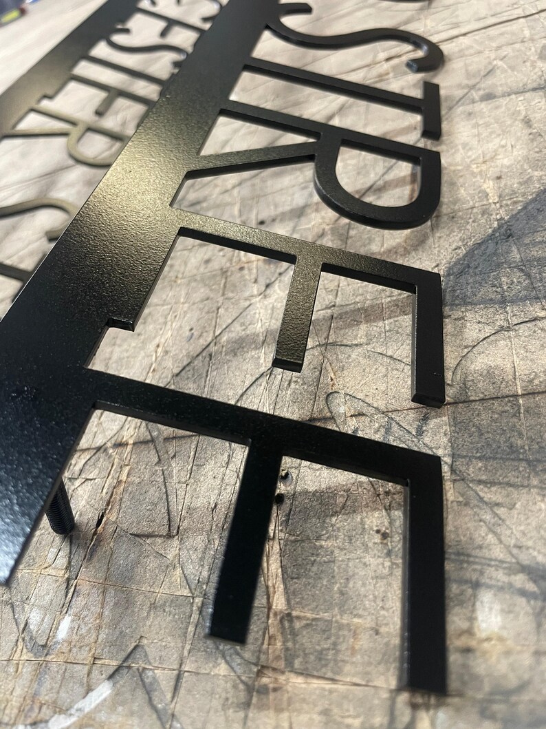 A close-up, low-angle view of the black metal sign, showing the laser-cut letters "STREET" with a slightly textured matte finish, on a gray surface.