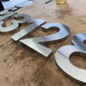 Brushed metal numbers "1328" are shown at an angle on a textured wooden surface, with the "2" and "8" in focus.