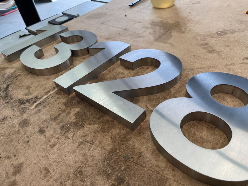 Brushed metal numbers "1328" are shown at an angle on a textured wooden surface, with the "2" and "8" in focus.
