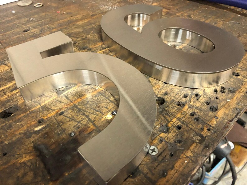 Two brushed metal numbers, a "5" and a "6", rest on a dark, scratched wooden surface, viewed from an angle.