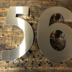 Two brushed metal numbers, a "5" and a "6", are placed side-by-side on a dark, scratched wooden surface.