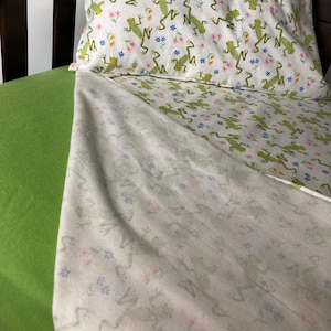 May include: A white pillowcase and sheet set with a repeating pattern of green frogs and small pink and blue flowers. The bed also has a solid lime green sheet. The bedding is on a dark brown wooden bed frame.