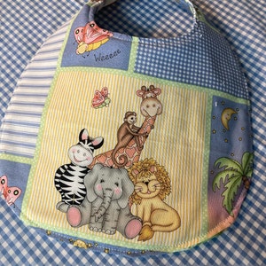 May include: A blue and yellow baby bib with a patchwork design featuring a giraffe, zebra, elephant, lion, and butterflies. The bib has the word "Weeeee" printed on it.