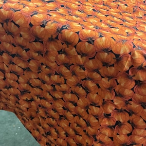 May include: A fabric featuring a repeating pattern of orange pumpkins with brown stems on a black background. A yellow measuring tape is visible in the upper right corner, showing a measurement of approximately 55 to 56 inches on the fabric edge.
