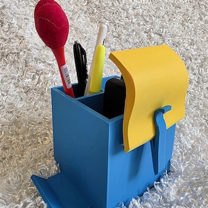 May include: A blue desk organizer with a built-in phone stand. It holds a red pen, black marker, yellow highlighter, and a pad of yellow sticky notes. The organizer is made of a solid material and sits on a white, textured surface.